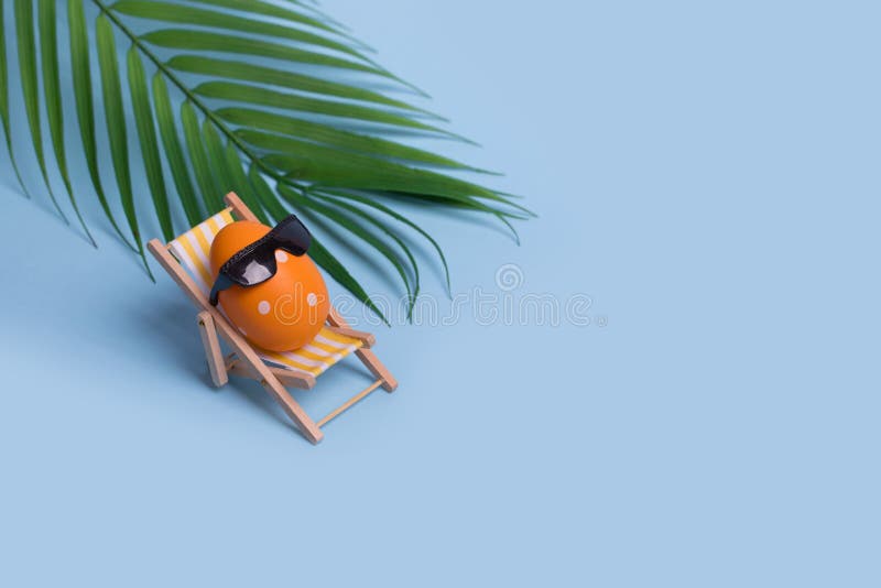 Creative funny composition with Easter egg with sunglasses sitting on deck chair and palm leaf on bright blue background royalty free stock photography
