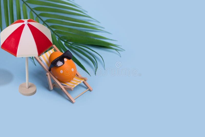 Creative funny composition with Easter egg with sunglasses sitting on deck chair and sun umbrella on bright blue background royalty free stock image