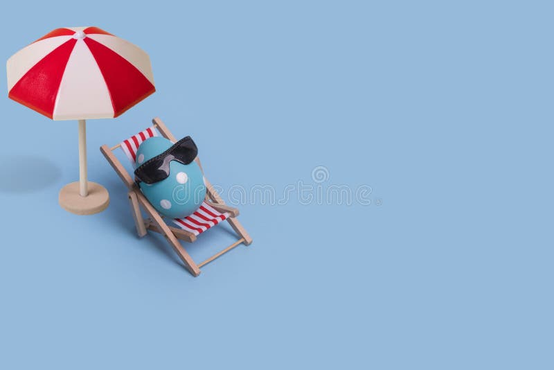 Creative funny idea with Easter egg with sunglasses while sitting on deck chair and sun umbrella on bright blue background stock image