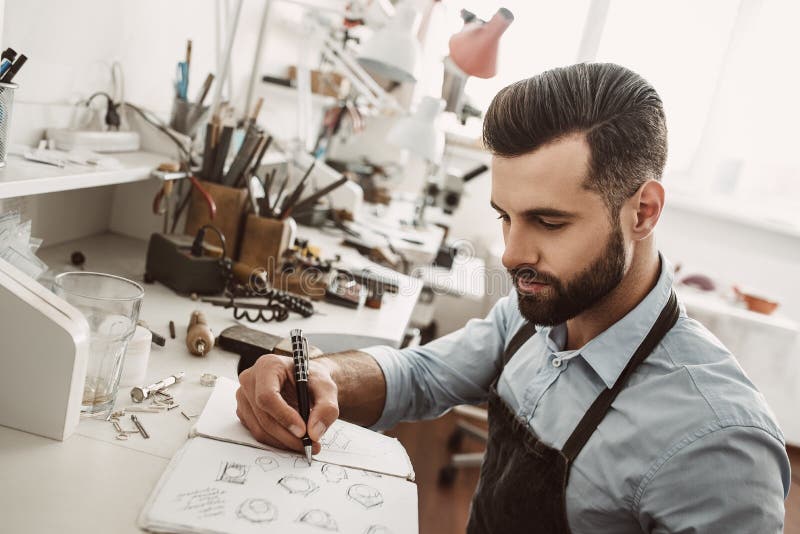 Creative work. Portrait of young bearded jeweler drawing a sketch of a new ring stock image Creative work. Portrait of young bearded jeweler drawing a sketch of a new ring stock image