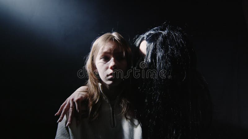 A creepy woman with black hair leans over to the girl and whispers in her ear
