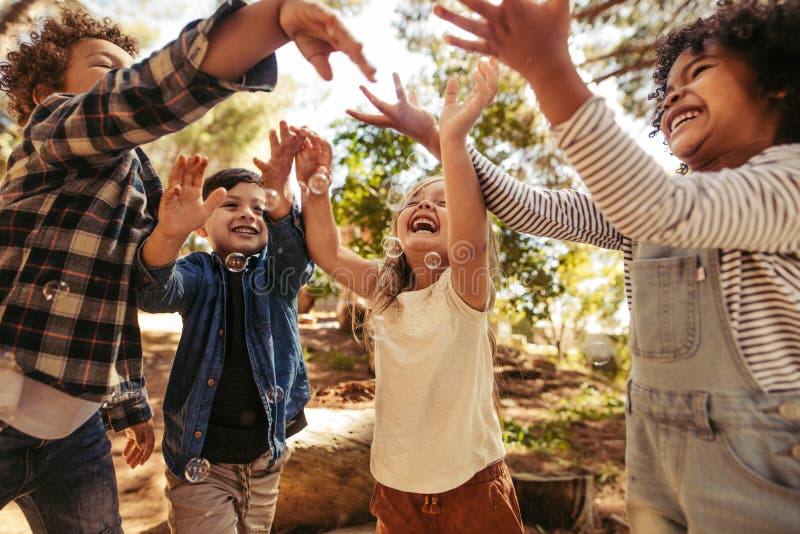 Group of kids playing with soap bubbles in forest. Boy blowing soap bubbles with friends trying to catch the bubbles. Group of kids playing with soap bubbles in forest. Boy blowing soap bubbles with friends trying to catch the bubbles