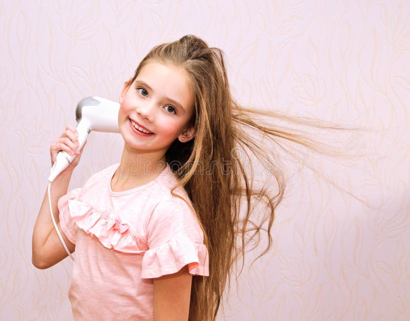 Criança de sorriso bonito da menina que seca seu cabelo longo com secador de cabelo foto de stock Criança de sorriso bonito da menina que seca seu cabelo longo com secador de cabelo foto de stock