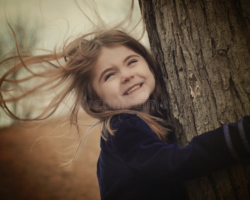 A little child is holding a tree with strong wind blowing in her hair. She is smiling and happy. Use for a weather or freedom concept. A little child is holding a tree with strong wind blowing in her hair. She is smiling and happy. Use for a weather or freedom concept.