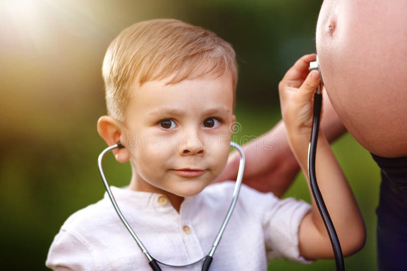 The child listens to the pregnant mother`s stomach with a stethoscope and looks at the camera. Portrait of a small boy in close-up. The child listens to the pregnant mother`s stomach with a stethoscope and looks at the camera. Portrait of a small boy in close-up