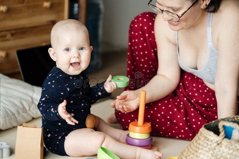 A criança pequena senta-se no assoalho e joga-se alegremente com os brinquedos coloridos com sua mãe no interior fotografia de stock royalty free A criança pequena senta-se no assoalho e joga-se alegremente com os brinquedos coloridos com sua mãe no interior fotografia de stock royalty free