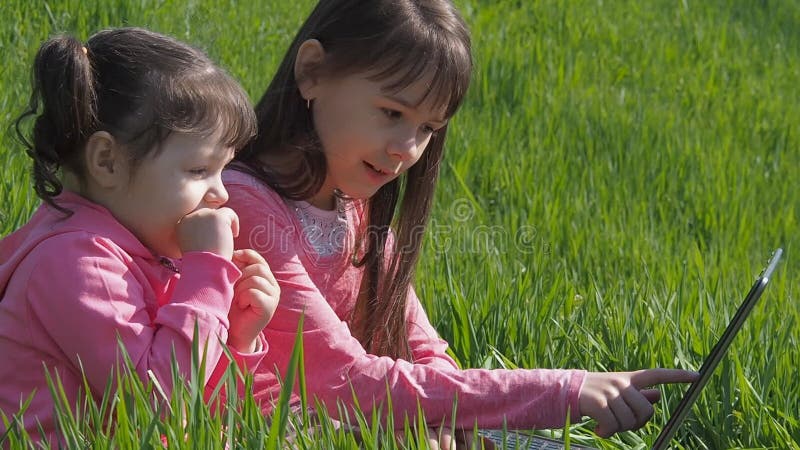 Crianças ao ar livre com portátil Meninas na grama verde Irmãs com um portátil no parque As crianças na roupa cor-de-rosa são pla