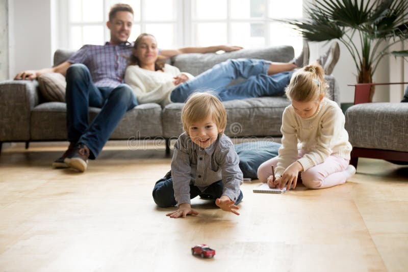 Crianças que jogam no assoalho, pais que relaxam no sofá em casa