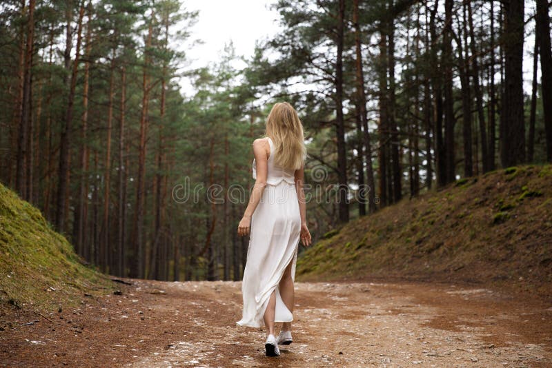 Crisalide bionda giovane bella di camminata della foresta della donna in vestito bianco in legno sempreverde fotografia stock Crisalide bionda giovane bella di camminata della foresta della donna in vestito bianco in legno sempreverde fotografia stock