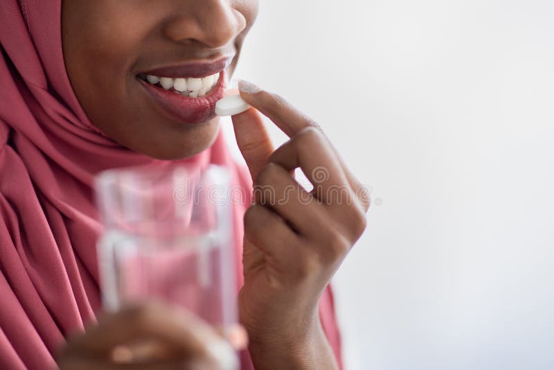 Black Muslim Woman In Hijab Holding Glass Of Water And Taking Pill