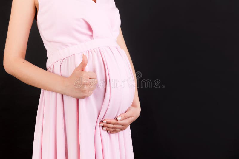 Cropped shot of positive pregnant woman in pink dress showing thumb up cool gesture over her baby bump at black background. Happy