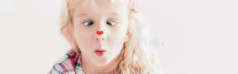 Cross-eyed kid looking at red heart sticker on her nose. Funny hilarious white Caucasian cute adorable child girl with blue eyes.