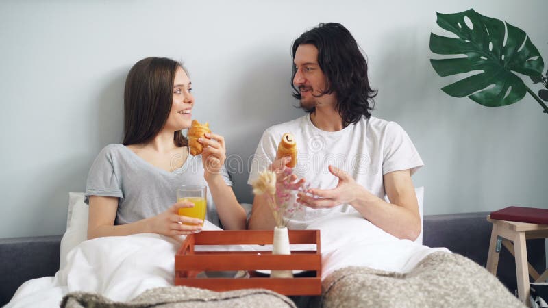 Cruasanes eaing de los pares alegres en la cama que charla y que ríe disfrutando de mañana