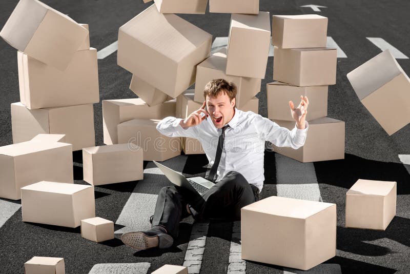 Crying and shocked businessman speaking phone and sitting with laptop on the asphalt road surrounded by falling carton boxes