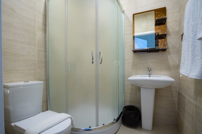 Cuarto de baño beige con la ducha esmaltada, espejo sobre el fregadero, toalla y retrete fotografía de archivo Cuarto de baño beige con la ducha esmaltada, espejo sobre el fregadero, toalla y retrete fotografía de archivo