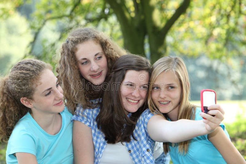 Four attractive teenage girls in a park posing close together and smiling for a self-portrait as one girl takes a photo on her mobile phone. Four attractive teenage girls in a park posing close together and smiling for a self-portrait as one girl takes a photo on her mobile phone