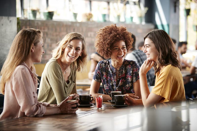 Cuatro amigos femeninos jovenes que hacen frente a la tienda y a la charla de Sit At Table In Coffee