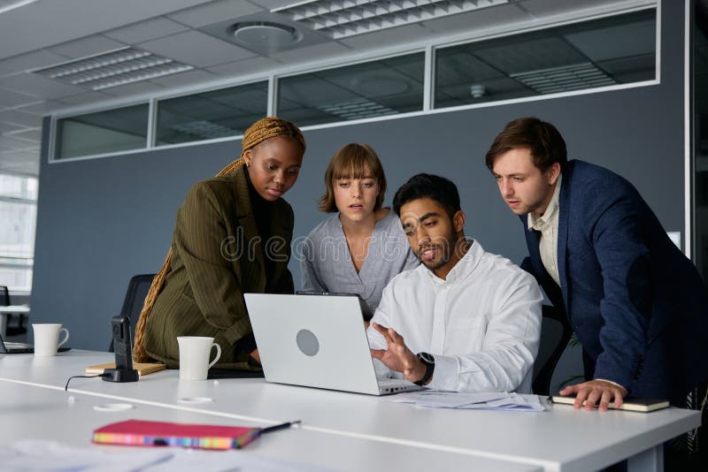 Four young adults wearing businesswear talking and gesturing by laptop on desk in corporate office. Four young adults wearing businesswear talking and gesturing by laptop on desk in corporate office