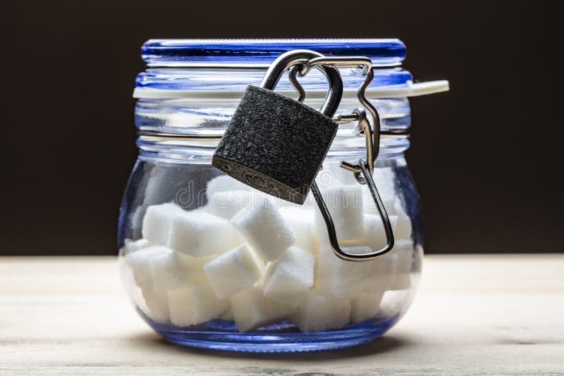 Cubes of white lump sugar in a glass jar with a lid closed with a lock, stands on a wooden table, on a black background. The concept of banning harmful foods in order to avoid diabetes. Cubes of white lump sugar in a glass jar with a lid closed with a lock, stands on a wooden table, on a black background. The concept of banning harmful foods in order to avoid diabetes