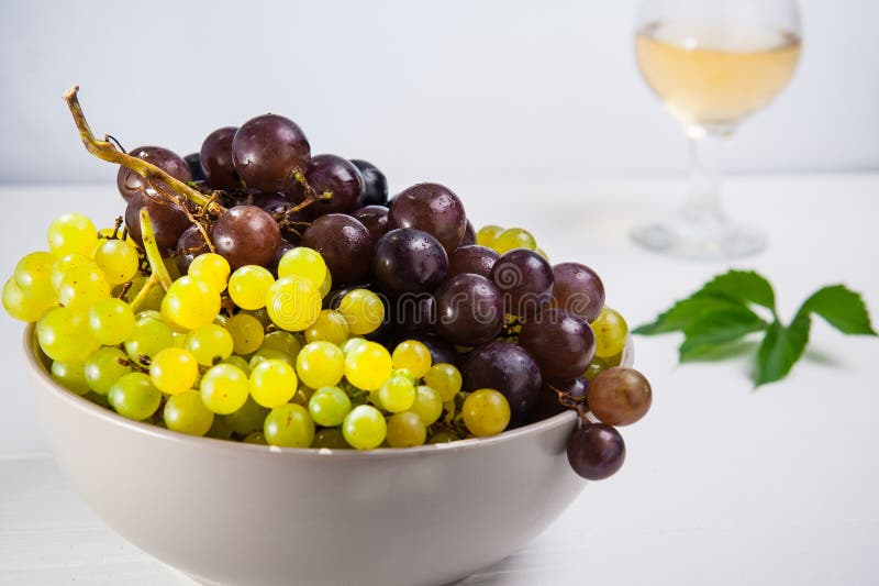 Cuenco de diversas uvas: bayas rojas, blancas y negras en la tabla de madera blanca con el vidrio del vino blanco y de la hoja ve imagen de archivo libre de regalías Cuenco de diversas uvas: bayas rojas, blancas y negras en la tabla de madera blanca con el vidrio del vino blanco y de la hoja ve imagen de archivo libre de regalías