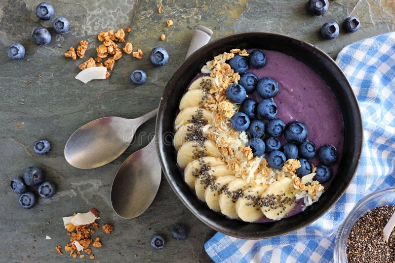 Cuenco del smoothie del arándano con el coco, los plátanos, las semillas del chia y el granola, sobre la opinión sobre un fondo o imagen de archivo libre de regalías Cuenco del smoothie del arándano con el coco, los plátanos, las semillas del chia y el granola, sobre la opinión sobre un fondo o imagen de archivo libre de regalías