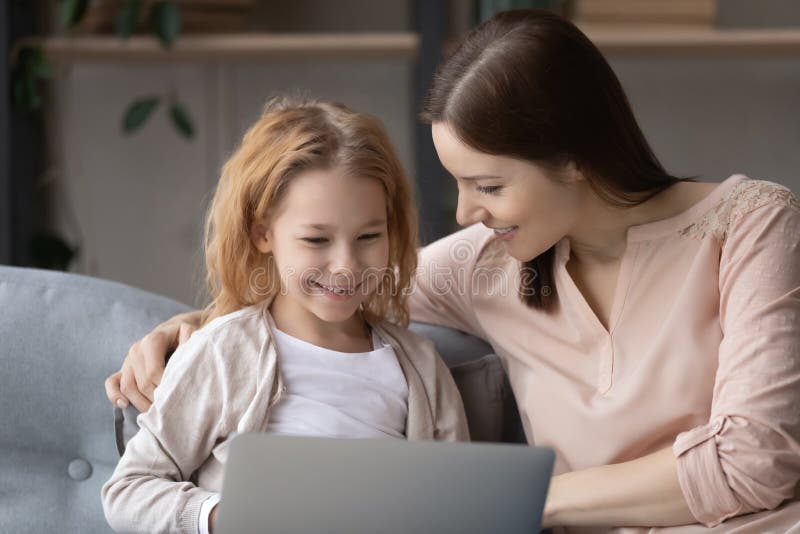 Cuidadosa joven madre enseñando a su pequeña hija usando aplicaciones de software. Cuidadosa joven madre enseñando a la pequeña y linda hija usando aplicaciones de software de computadora apoyándose en un cómodo sofá en casa. adicto a la tecnología moderna uniendo a la familia de dos generaciones jugando en línea.