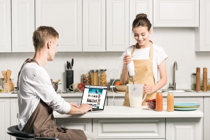 girlfriend cooking and boyfriend using laptop with loaded amazon page in kitchen. girlfriend cooking and boyfriend using laptop with loaded amazon page in kitchen