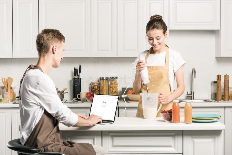 girlfriend cooking and boyfriend using laptop with loaded google page in kitchen. girlfriend cooking and boyfriend using laptop with loaded google page in kitchen