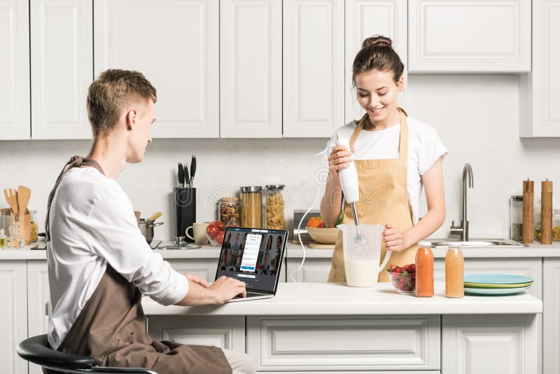 girlfriend cooking and boyfriend using laptop with loaded linkedin page in kitchen. girlfriend cooking and boyfriend using laptop with loaded linkedin page in kitchen