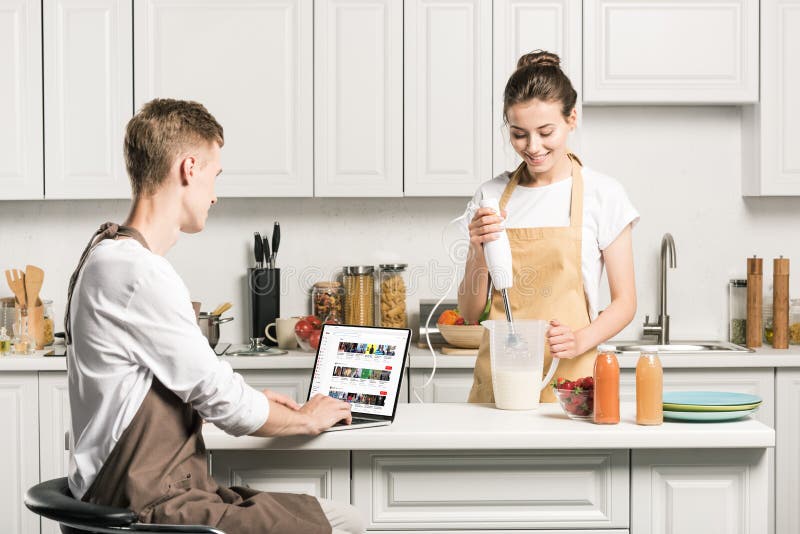 girlfriend cooking and boyfriend using laptop with loaded youtube page in kitchen. girlfriend cooking and boyfriend using laptop with loaded youtube page in kitchen