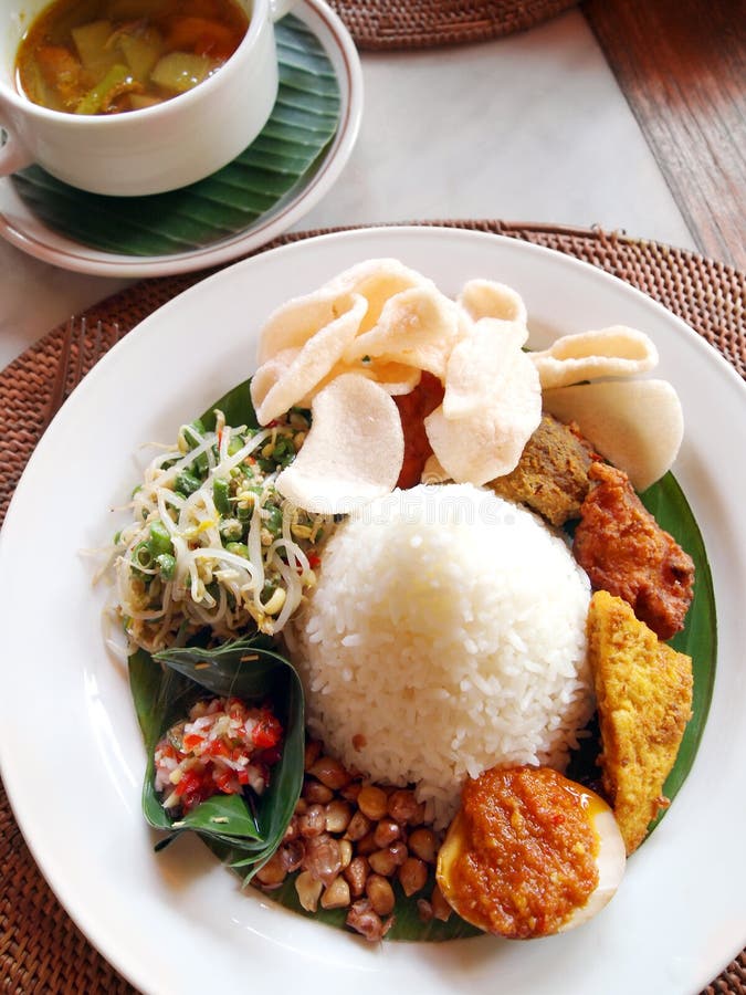 A photograph picture of a set meal of traditional indonesia cooking popular in Java, Sumatra and Bali. Nasi campur, or mixed assorted dishes with steamed white rice. Dishes include hot spicy rendang curry meats, fish, tempeh fermented beans, egg and local vegetables. Served with daily soup, prawn crackers, peanuts and sambal chilli. Delicious tasty cuisines, popular main course item with locals and tourists. Vertical color format photo. Beautiful professional foods photography with natural day lighting. A photograph picture of a set meal of traditional indonesia cooking popular in Java, Sumatra and Bali. Nasi campur, or mixed assorted dishes with steamed white rice. Dishes include hot spicy rendang curry meats, fish, tempeh fermented beans, egg and local vegetables. Served with daily soup, prawn crackers, peanuts and sambal chilli. Delicious tasty cuisines, popular main course item with locals and tourists. Vertical color format photo. Beautiful professional foods photography with natural day lighting.