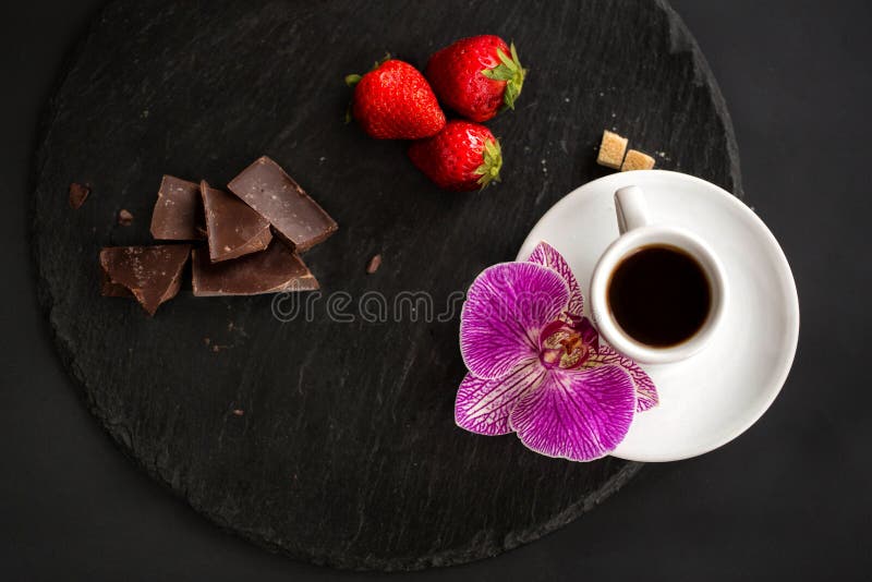 Cup of black coffee, brown sugar, strawberries, black chocolate on a round slate on a black background