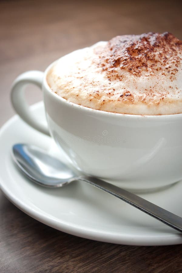 A cup of Cappuccino with a spoon on a brown table