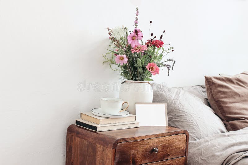 Cup of coffee and books on retro wooden bedside table. Blank greeting card mockup. White ceramic vase. Buquet of cosmos