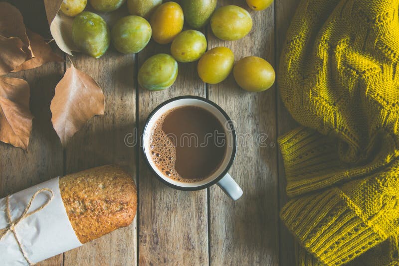 Cup of hot cocoa, whole grain rye bun, scattered yellow and green plums in craft paper bag. Dry leaves knitted sweater, fall