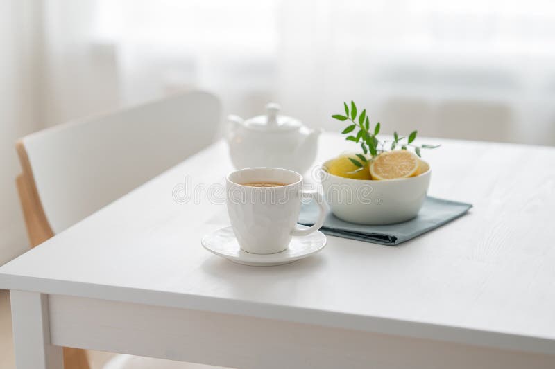 A cup of tea with lemon and a teapot on a white table against the background of a kitchen window