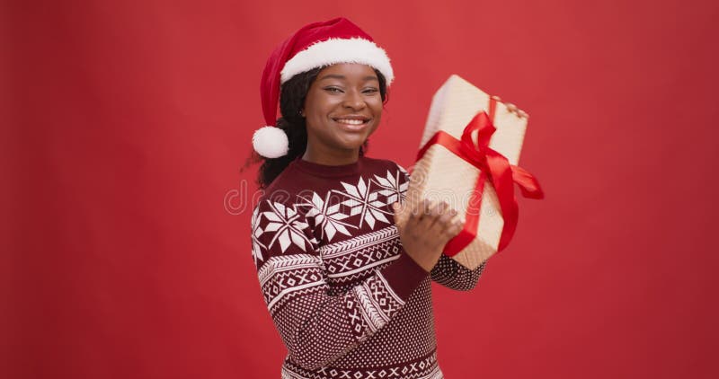 Curious black lady in santa hat shaking gift box, enjoying Christmas holiday, red background