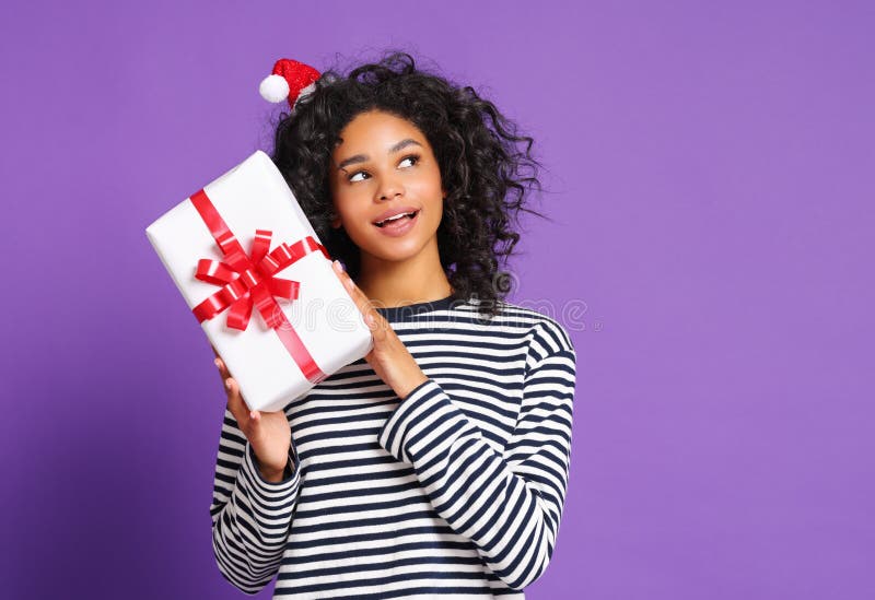 Curious smiled  ethnic woman  with santa hat   checking Christmas gift on colorful  purple  background