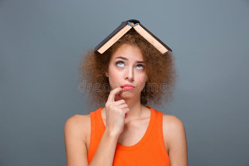 Portrait of curly annoyed bored tired young woman in orange top looking funny with book on head touching lips with forefinger and looking up. Portrait of curly annoyed bored tired young woman in orange top looking funny with book on head touching lips with forefinger and looking up