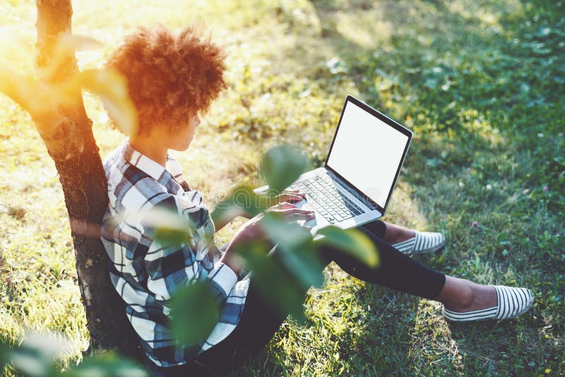 Rear view of cute black teenager girl in checkered shirt with laptop sitting on grass of spring meadow, charming mixed female student with curly afro hair and net-book is working while leaning on tree. Rear view of cute black teenager girl in checkered shirt with laptop sitting on grass of spring meadow, charming mixed female student with curly afro hair and net-book is working while leaning on tree