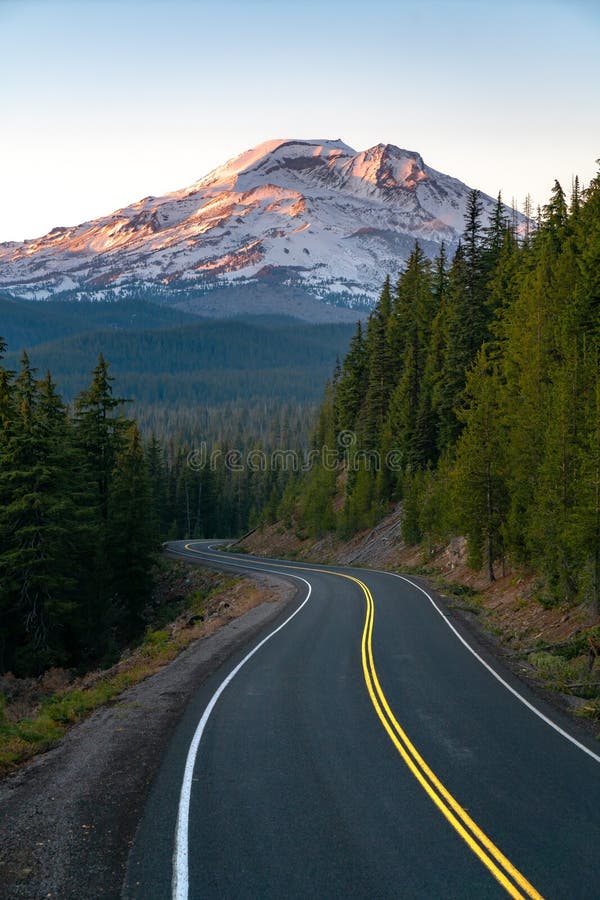 A Curvy Road Surrounded By Green Trees With High Rocky Snow Covered