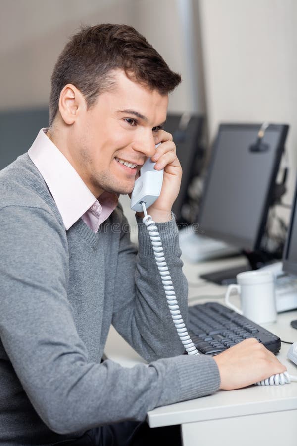 Smiling male customer service representative using landline phone at desk in office
