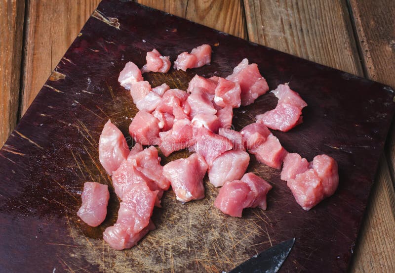 Cut fresh pork cubes on wooden board with knife.