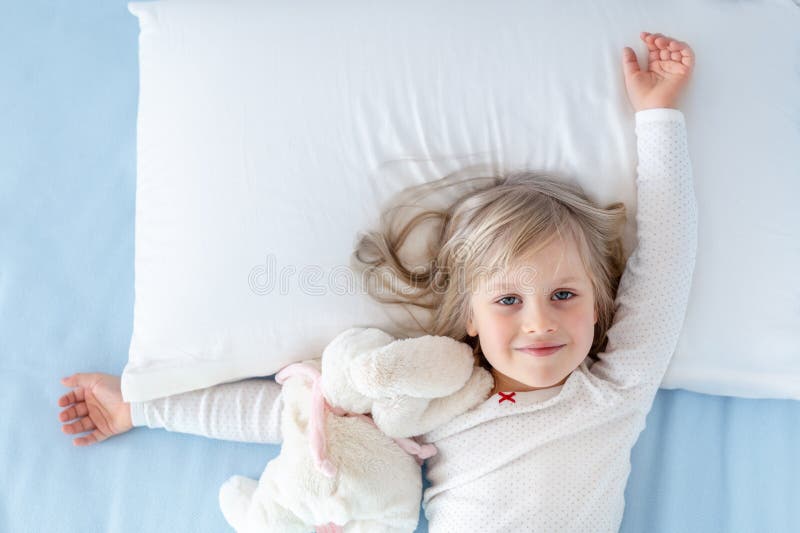 Cute adorable caucasian blond little toddler kid girl lying in bed on white pillow. Adorable sweet happy smiling child stretching