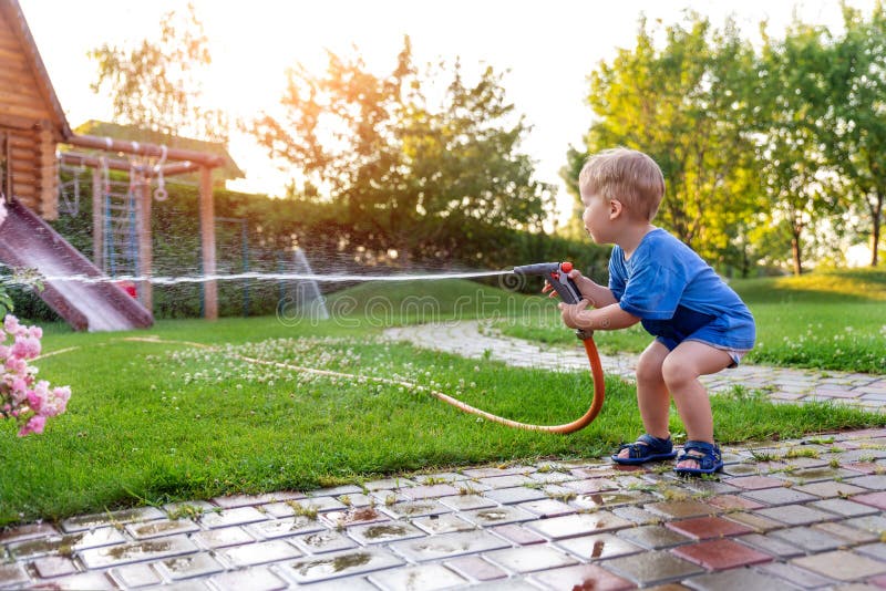 Cute adorable caucasian blond toddler boy enjoy having fun watering garden flower and lawn with hosepipe sprinkler at