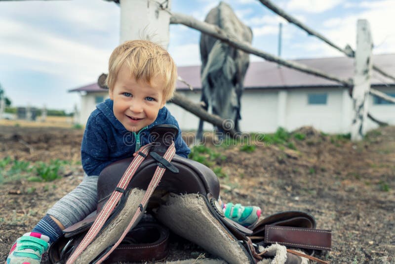 Cute adorable caucasian little smiling toddler portrait boy sitting on saddle on ground enjoy having fun riding supposed horse on countryside farm outdoors. Happy childhood on country ranch.