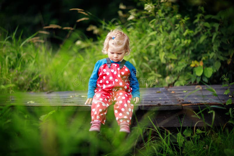 Cute adorable toddler girl sitting on wooden bridge and throwing small stones into a creek. Funny baby having fun with