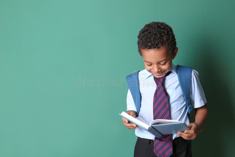 Cute African-American schoolboy reading book on color background