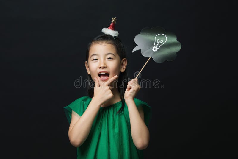 Cute asian girl child dressed in a green dress holding an idea bulb stick with a christmas head decoration on a black background.