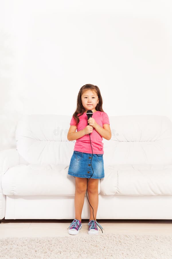 Cute little Asian five years old girl with dark long hair singing to microphone performing standing in living room. Cute little Asian five years old girl with dark long hair singing to microphone performing standing in living room